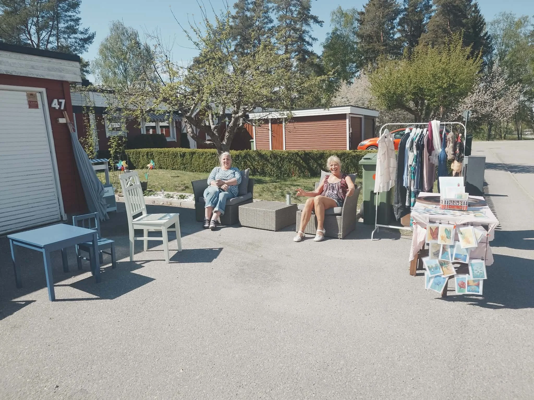 Två kvinnor som sitter utomhus på en för som antagligen är en bakgård eller trädgård. Det finns en klädställning med kläder, en liten blomkruka och en bricka med böcker eller vykort.