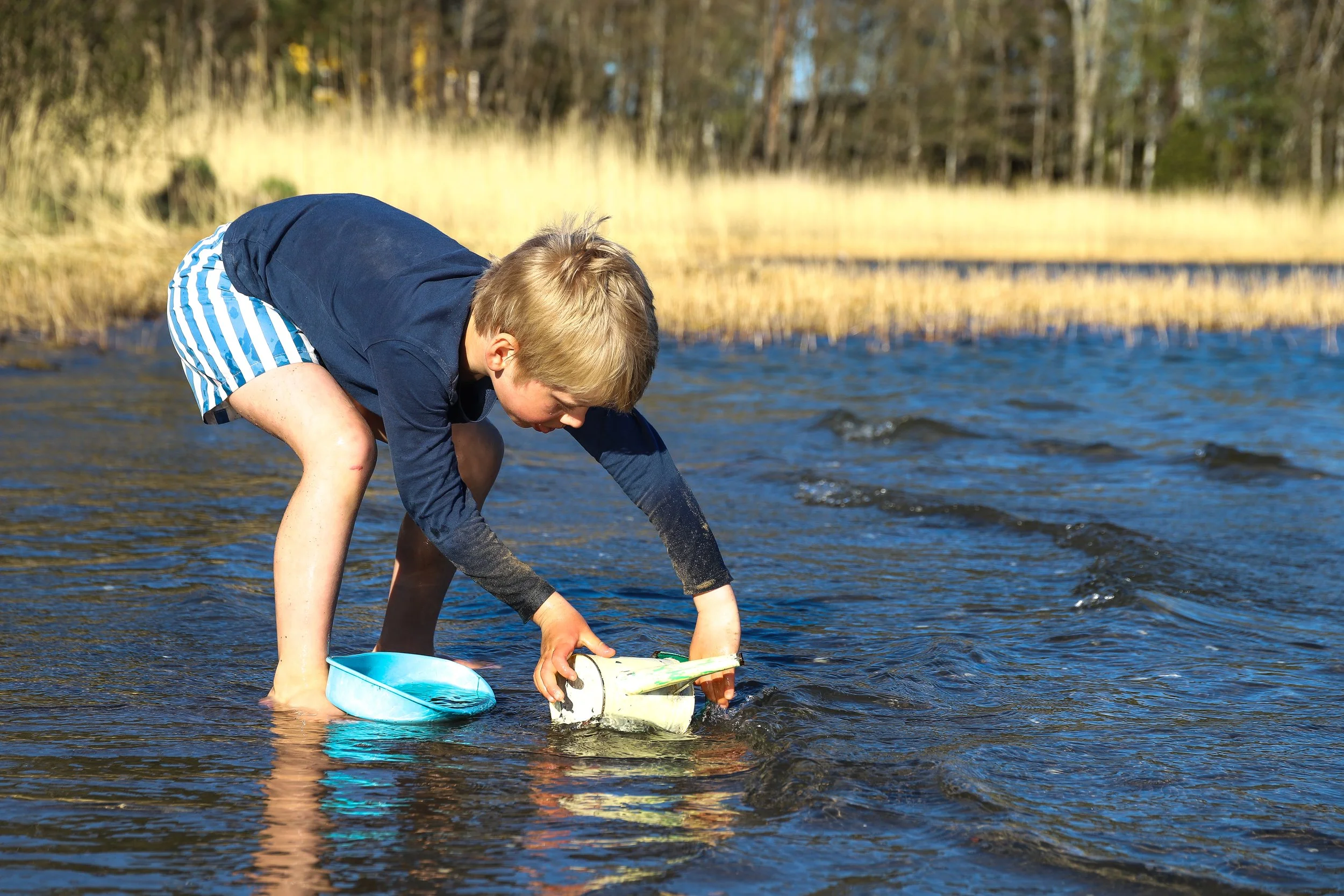 En pojke bär en blå jacka och blåvita badbyxor som står i vattnet, leker med en rullad plastflaska, omgiven av natur med träd och högt gräs i bakgrunden.