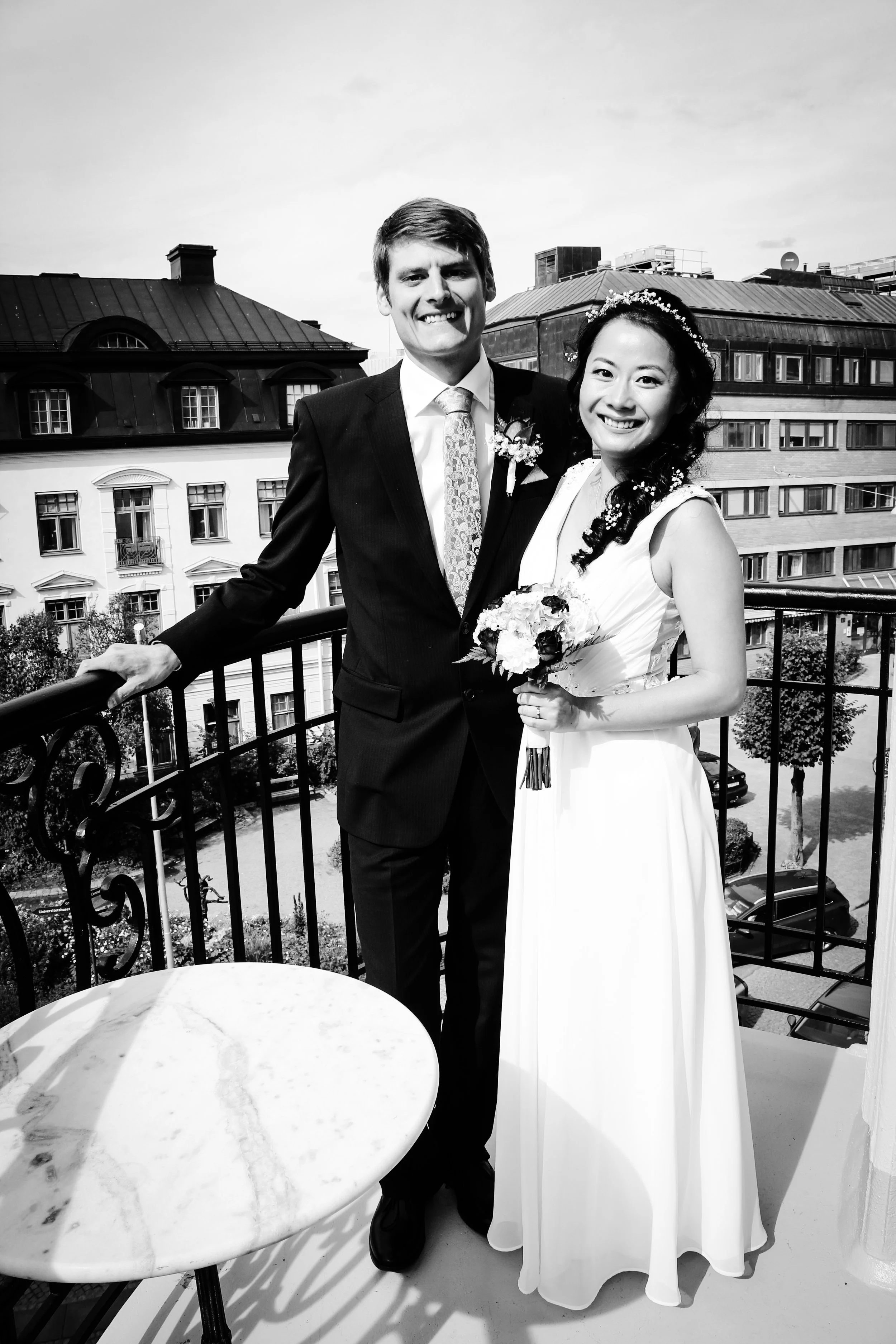 Ett brudpar på en balkong, ler och håller i en brudbukett, med stadshus i bakgrunden, svartvitt foto.