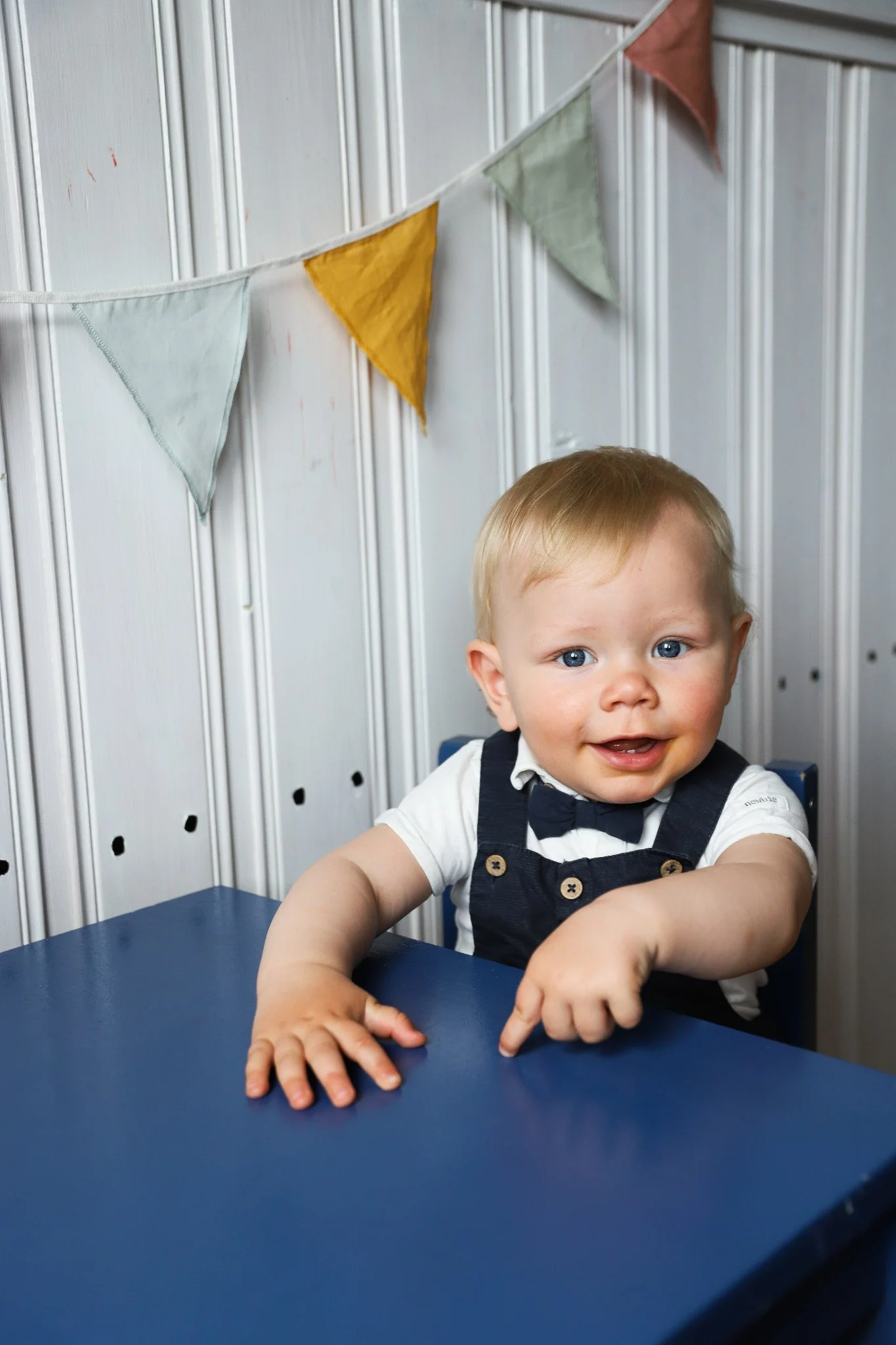 En ung pojke med ljus hud, blond hår och blå ögon, som ler och tittar mot kameran. Han sitter vid ett blått bord och är klädd i en vit skjorta och mörkblå hängselbyxor med knappar. Bakgrunden består av en vita vägg med vertikala ränder och ett festband av trekantiga tygflagor i mjuka färger.