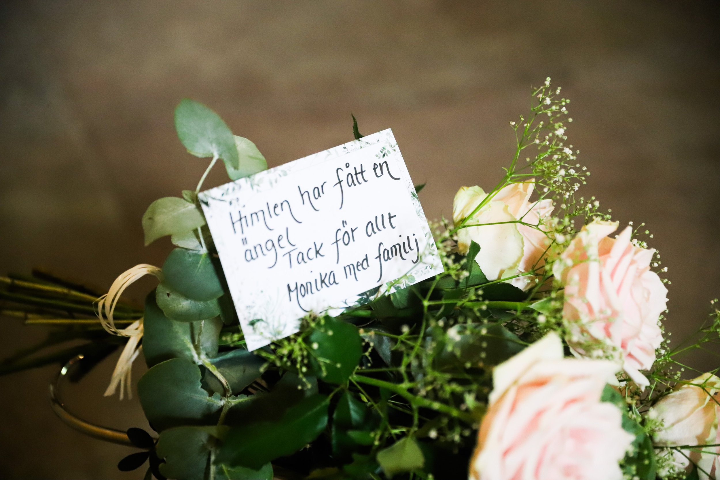 Blomsterarrangemang med en lapp som säger 'Himlen har fått en ängel. Tack för allt, med kärlek Monika med familj'.