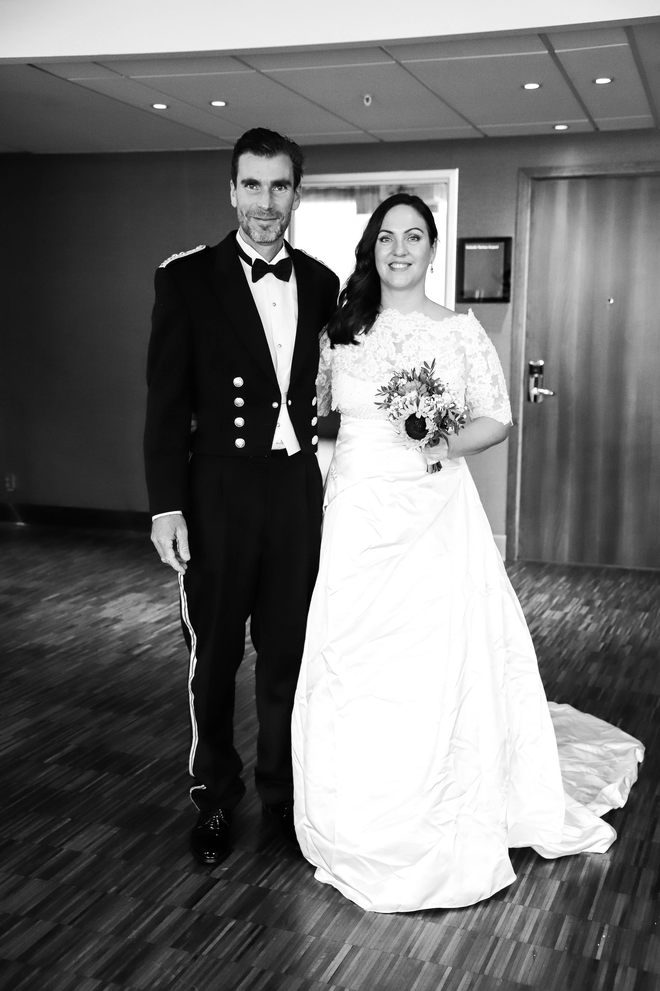En man i formell militäruniform och en kvinna i bröllopsklänning, båda står inomhus på ett bröllop, med en blomsterbukett i kvinnans hand.