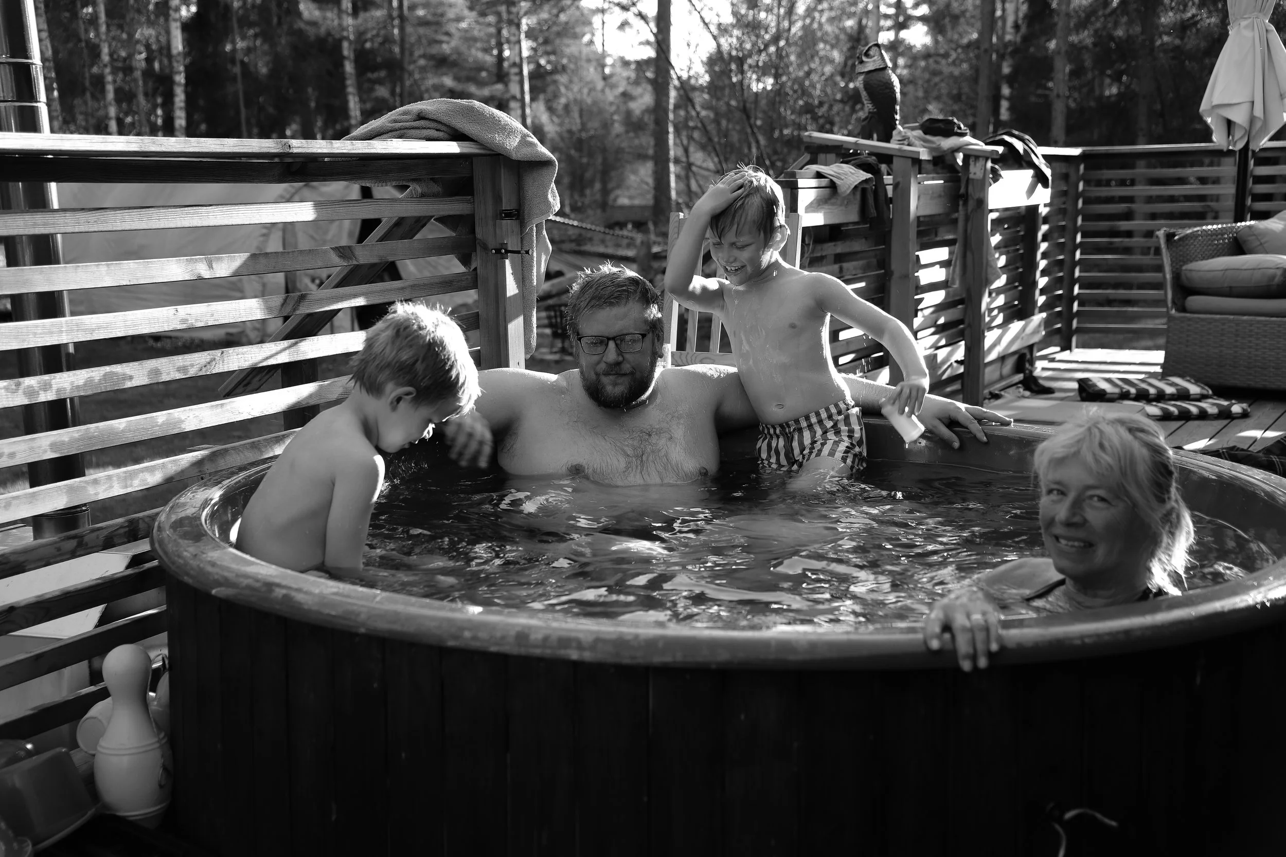 Familj som badar i ett utomhusbadkar, barn leker och har kul, solig dag, trädtak i bakgrunden, utomhusmiljö.