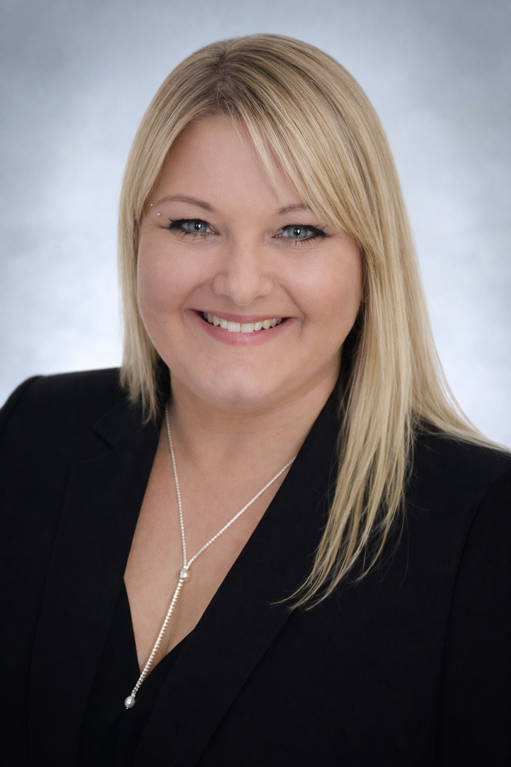 Portrait of a smiling woman with blonde hair, wearing a black blazer and a delicate necklace with pearls, against a gray background.