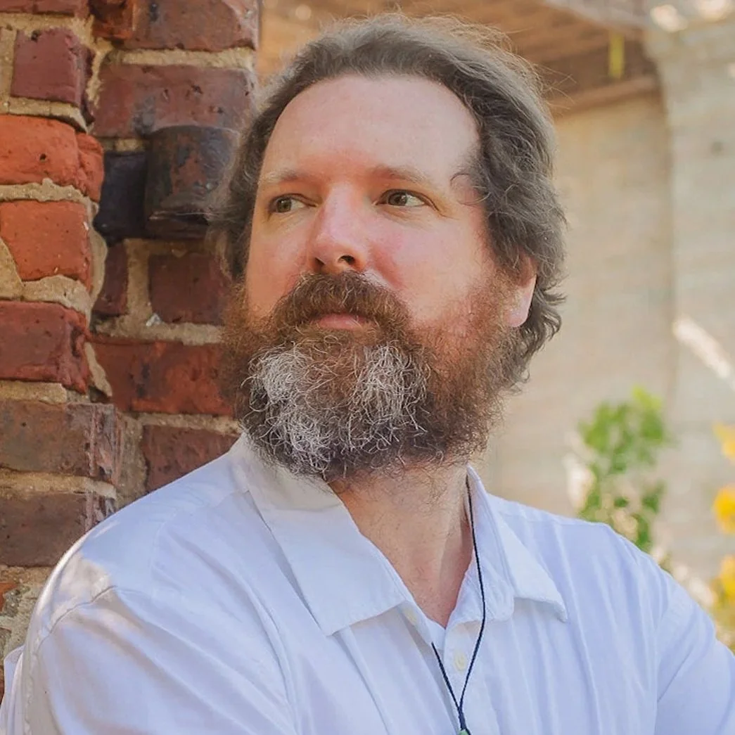 A man with a beard and mustache, wearing a white shirt, standing outdoors near a brick wall with a blurred background of trees and sunlight.