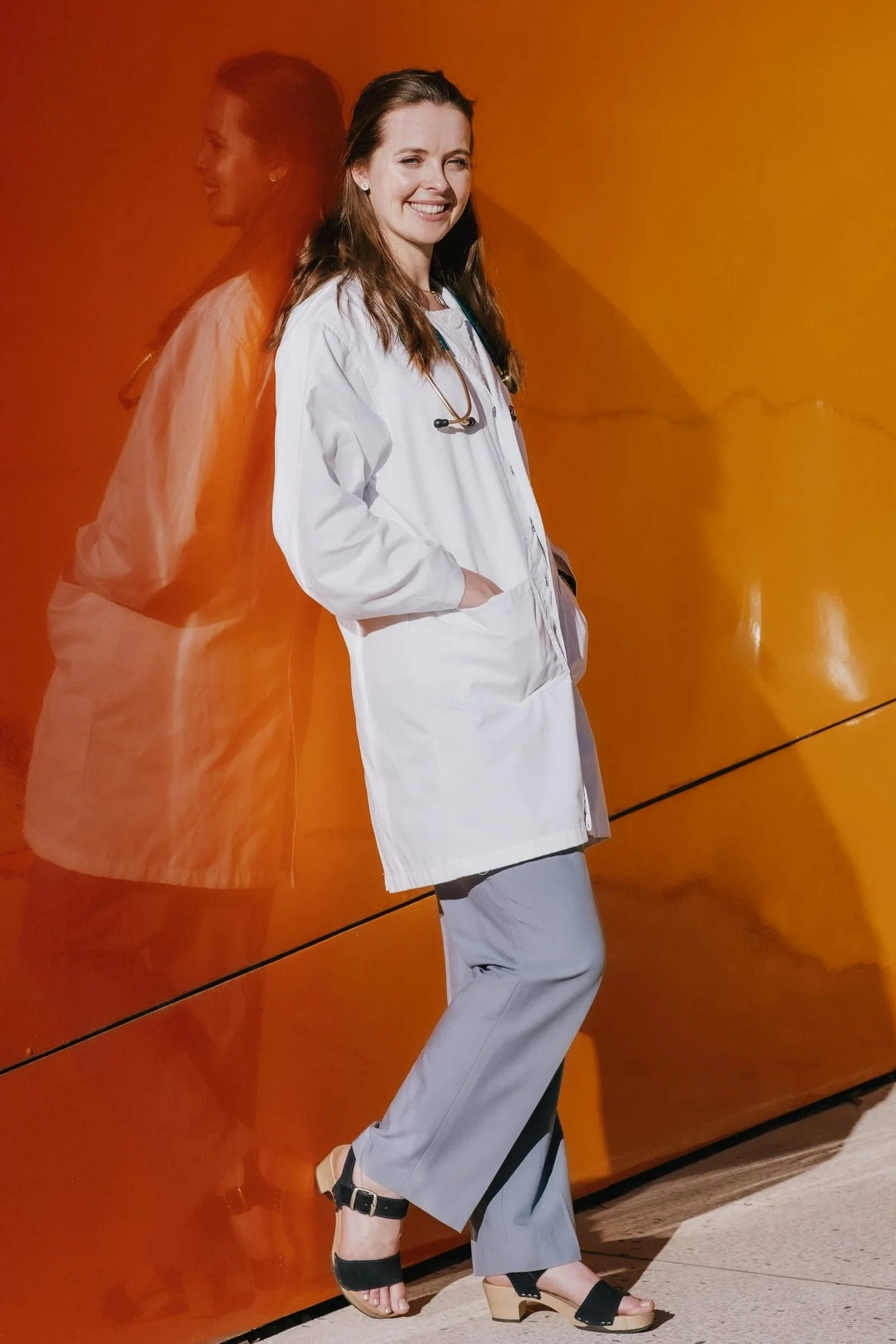 A woman wearing a white lab coat with a stethoscope around her neck, standing against an orange wall and smiling.
