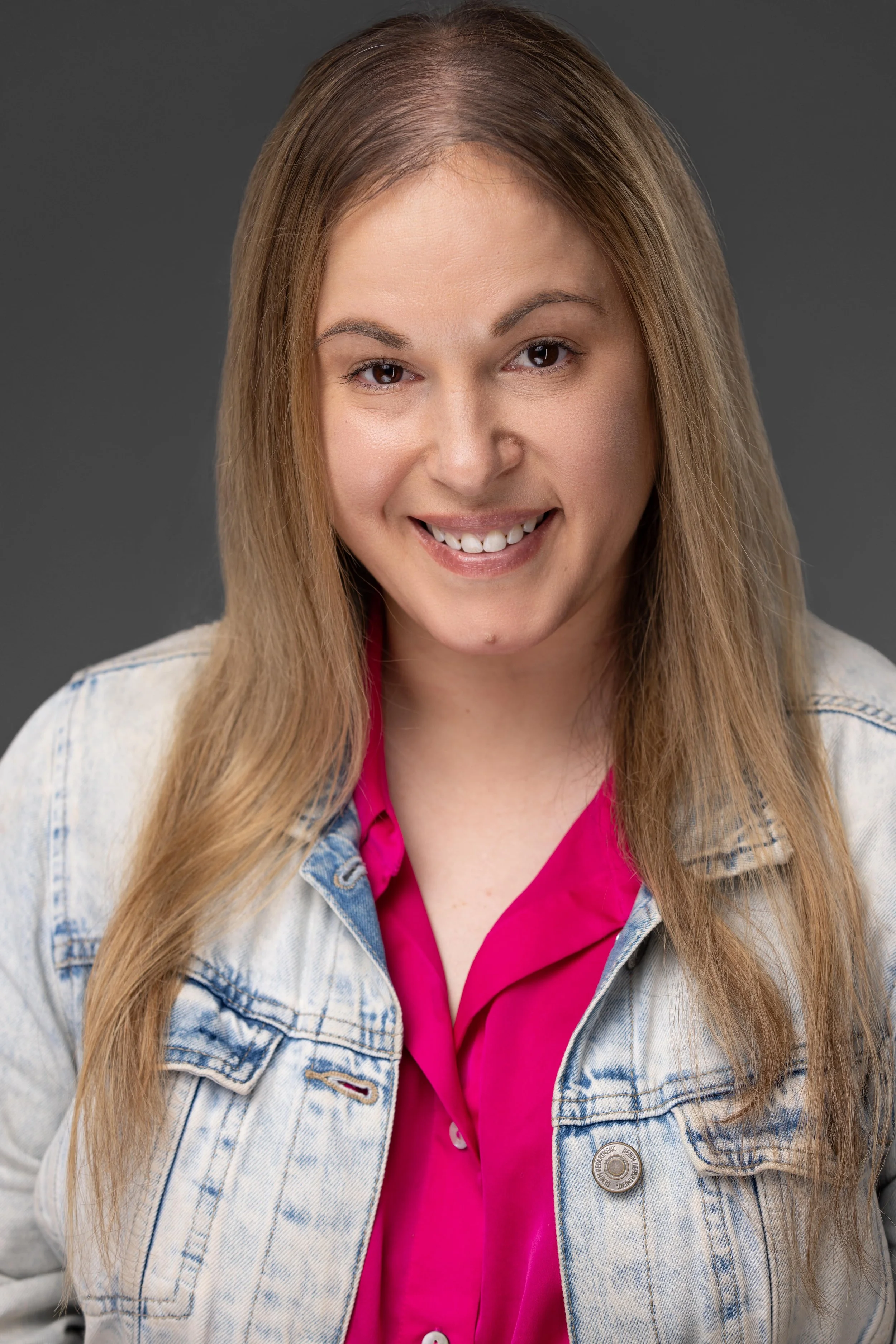 Close-up of a young woman with long, wavy light brown hair, smiling, wearing a light denim jacket over a bright pink shirt, against a neutral gray background.