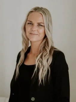 A woman with long blonde hair wearing a black blazer and black top, smiling softly against a light background.