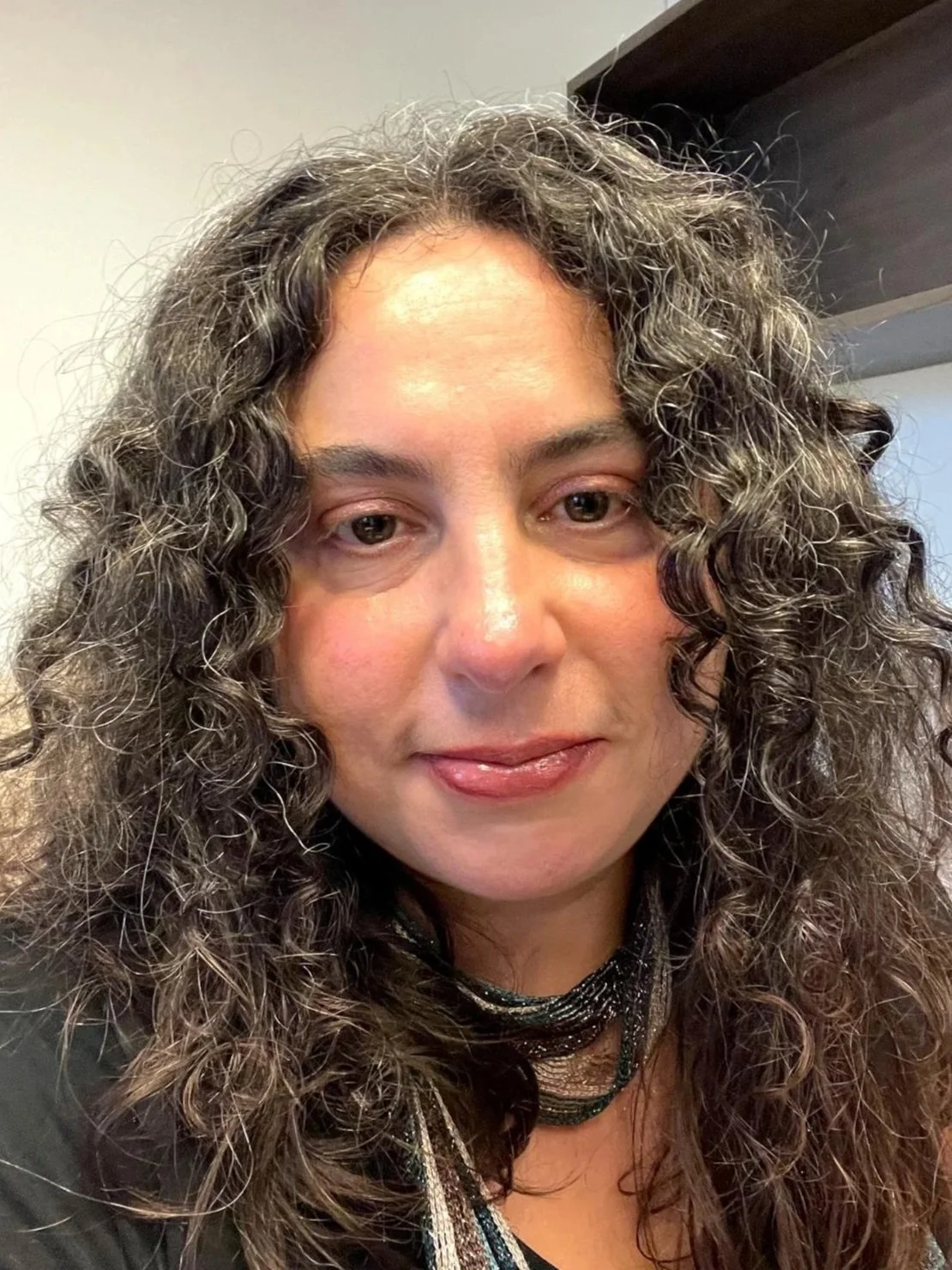 Close-up of a woman with curly, shoulder-length hair, wearing a black top and a multi-layered necklace, indoors with plain walls and a dark shelf behind her.