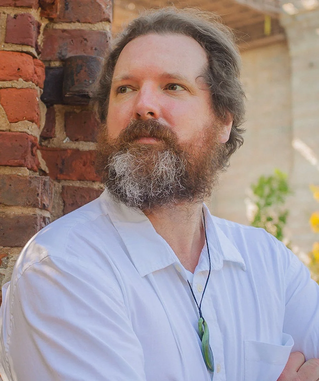 A bearded man with long hair wearing a white shirt and a pendant around his neck, standing outdoors next to a brick wall with greenery in the background.