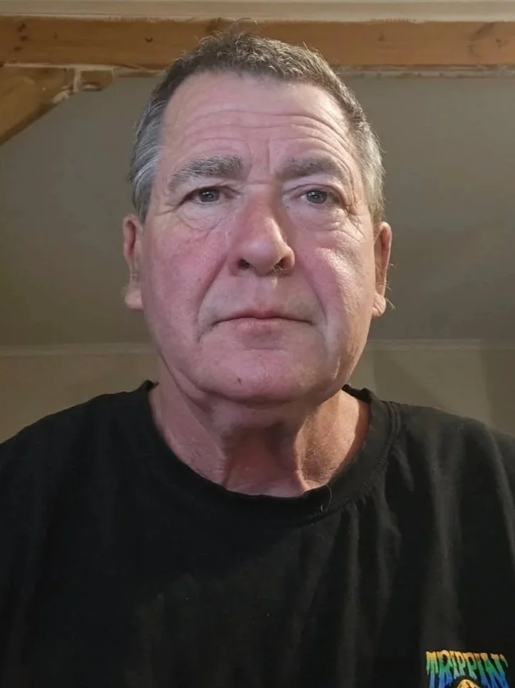 A middle-aged man with short gray hair, wearing a black T-shirt, looking directly at the camera in a room with a wooden ceiling beam.