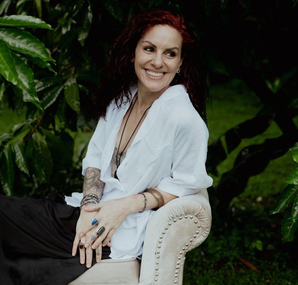 A woman with red hair and tattoos, sitting on a beige upholstered chair outdoors surrounded by green foliage, smiling and looking to the side.