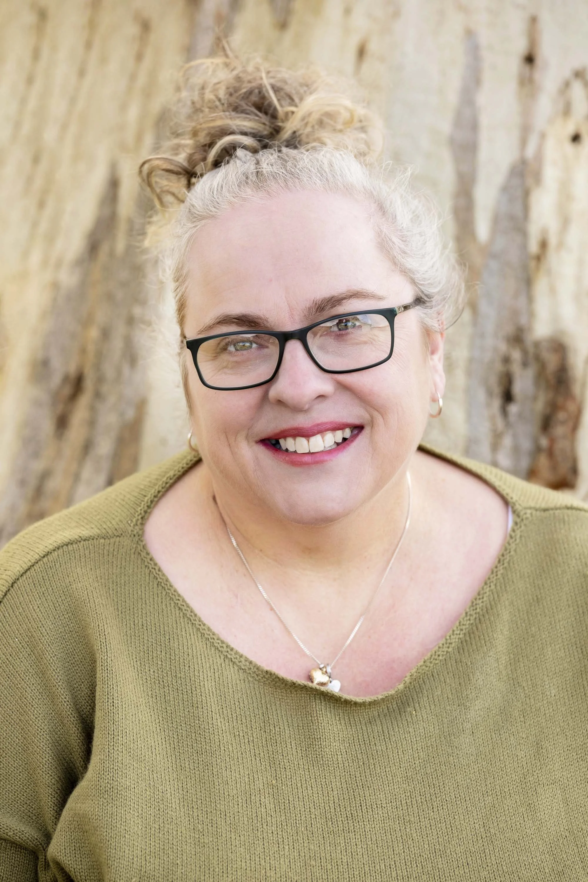 A smiling woman with curly blonde hair tied up, wearing glasses, a green sweater, and a necklace, standing outdoors with a large tree in the background.