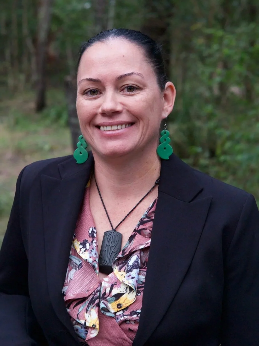A woman smiling outdoors, wearing a black blazer, floral blouse, green earrings, and a black pendant necklace, with a background of green trees.