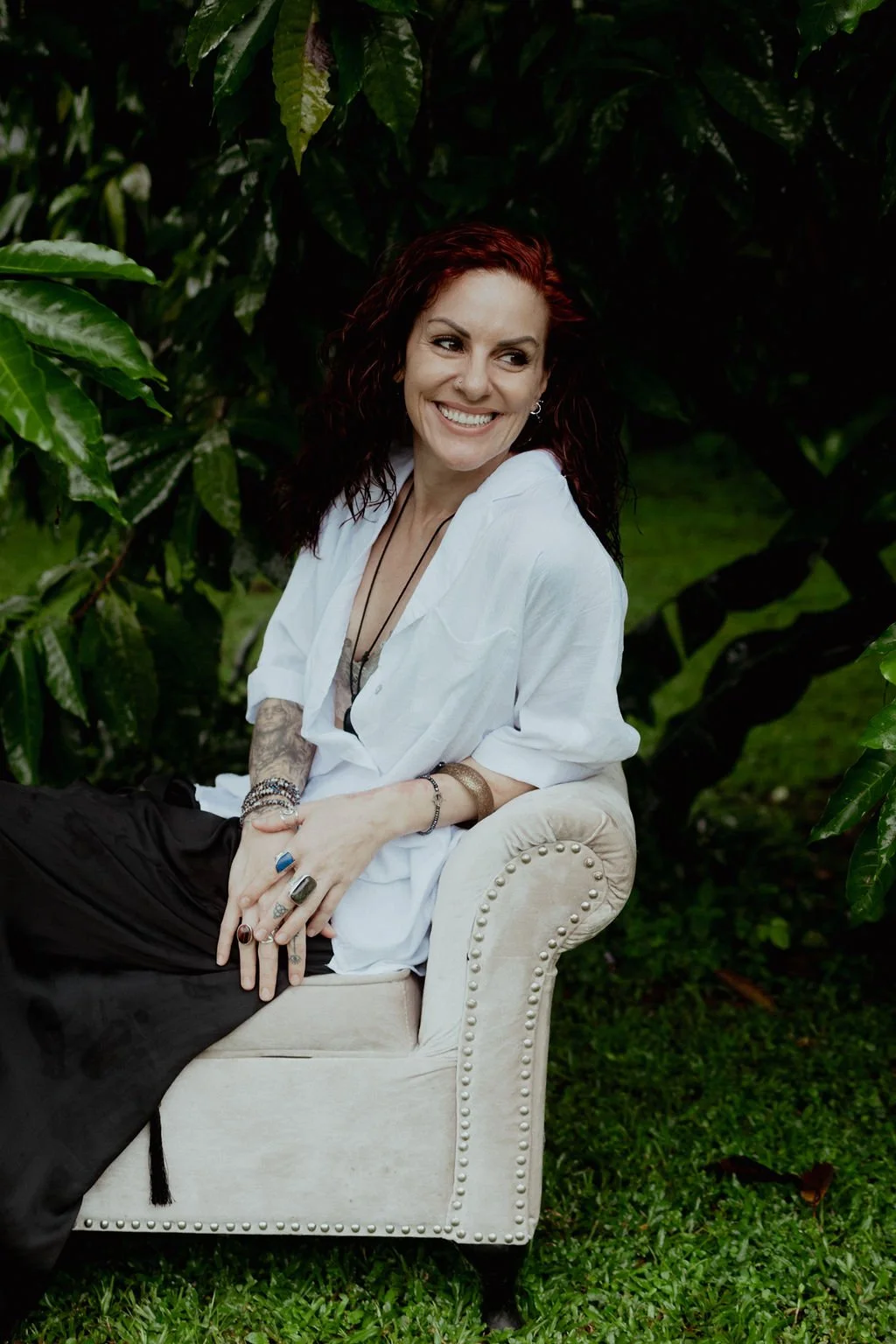 Woman with curly red hair sitting on a beige vintage armchair outdoors surrounded by green foliage, smiling and looking to the side.