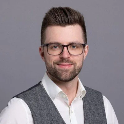 Portrait of a man with glasses, brown hair, beard, wearing a white shirt and gray vest against a gray background.
