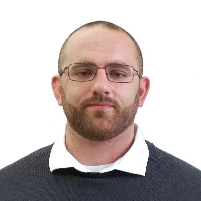 A man with glasses, a beard, wearing a dark sweater over a white shirt, posed against a plain white background.