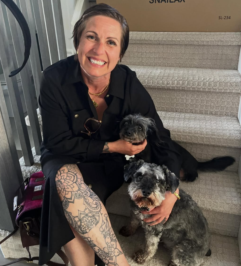 A woman with short dark hair, tattoos on her leg, and wearing a black outfit, sitting on stairs and smiling while holding two small dogs.