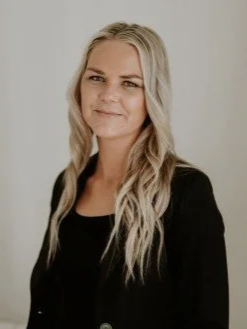 A woman with long, wavy blonde hair wearing a black blazer and black top, standing against a plain light-colored background, smiling gently.