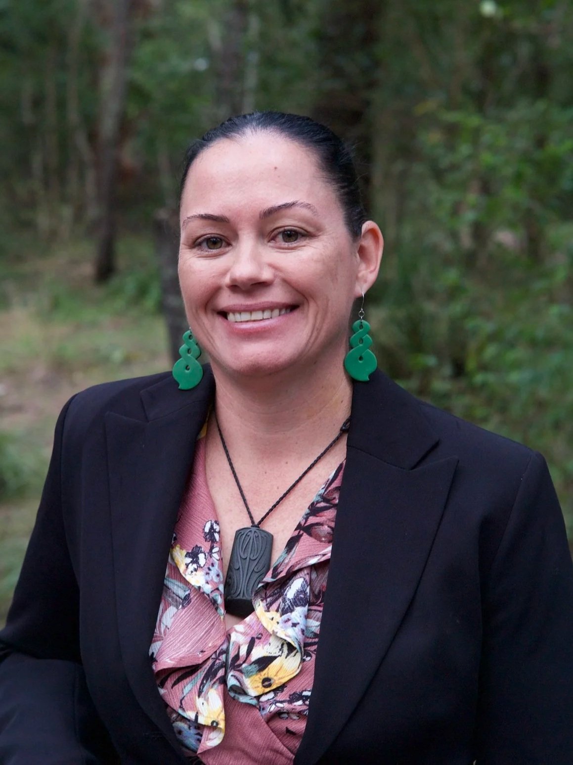 A woman with dark hair pulled back, smiling, outdoors with trees in the background, wearing a black blazer, floral blouse, large green earrings, and a black necklace with a carved pendant.