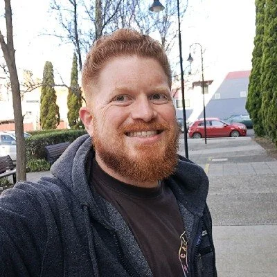 A man with red hair and a beard taking a selfie outdoors on a sunny day.