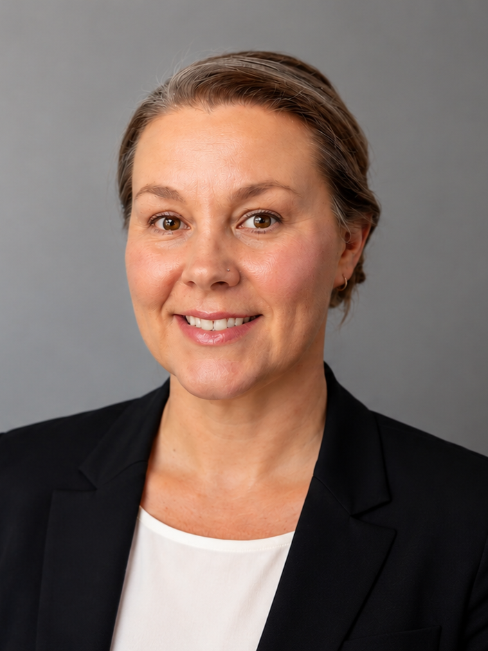 A professional woman with light skin, short brown hair with grey streaks, wearing a black blazer over a white top, smiling, against a plain grey background.