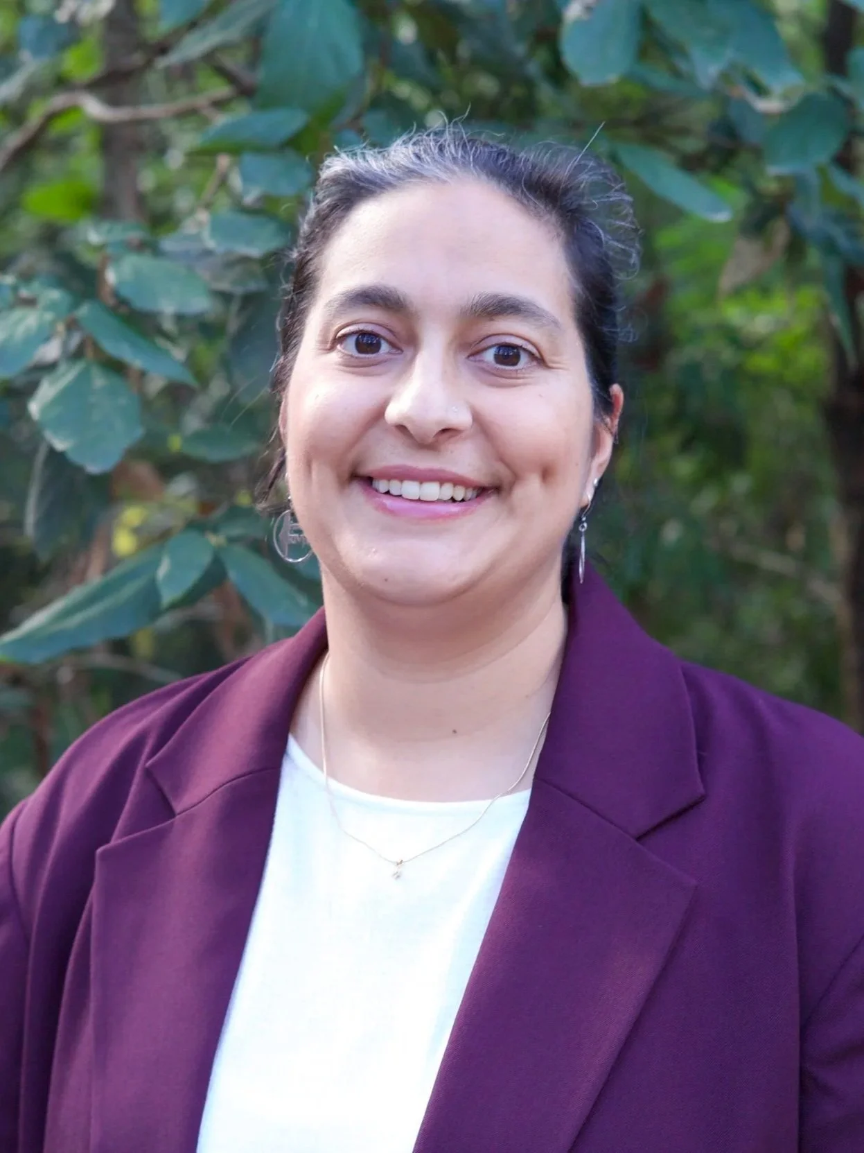A woman with dark hair tied back, smiling, wearing a purple blazer, white shirt, with greenery in the background.
