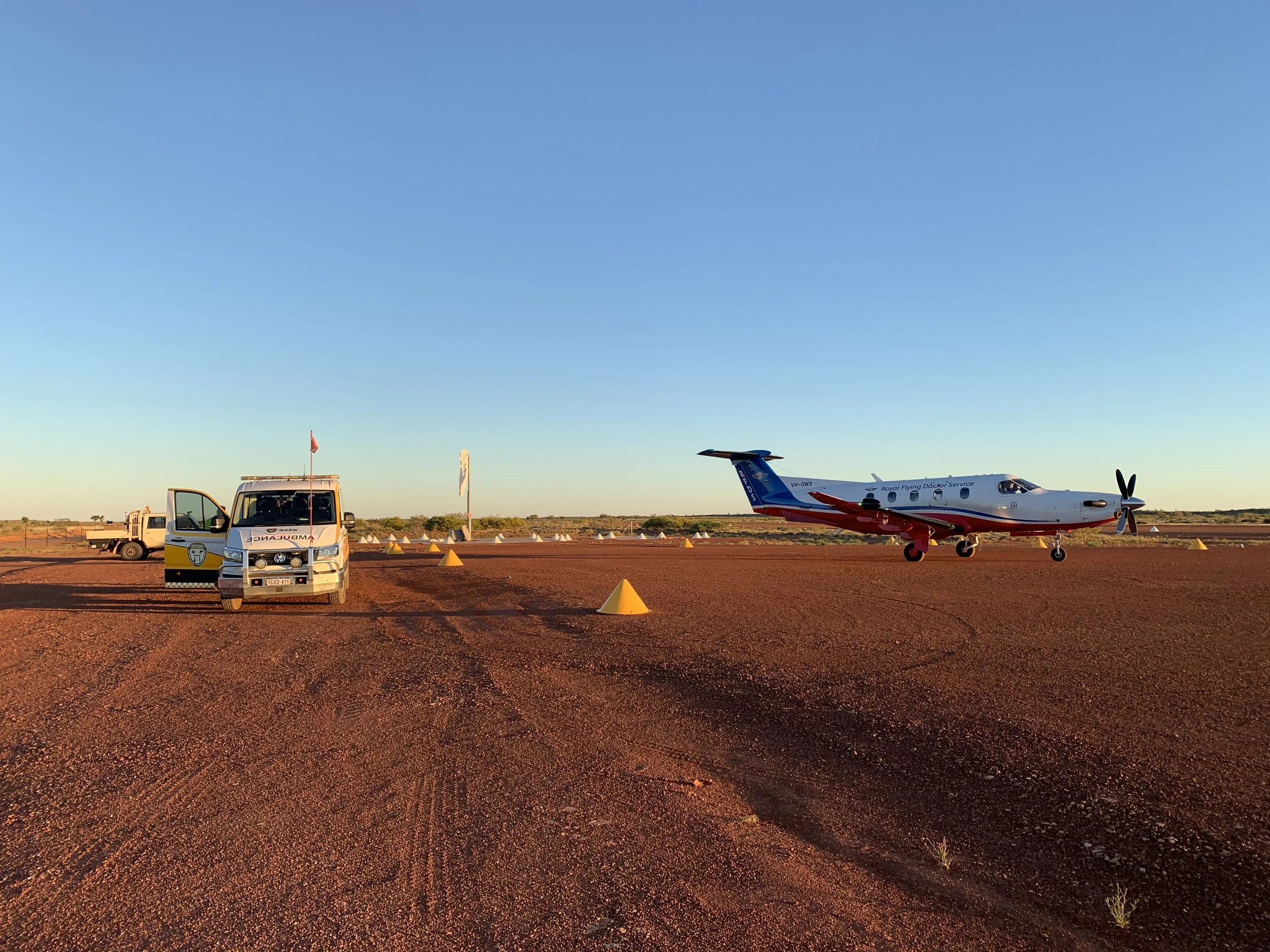 Small aircraft with Royal Flying Doctor Service logo parked on a dirt airstrip with yellow cones, a white vehicle, and flagpoles nearby under a clear blue sky at sunset.
