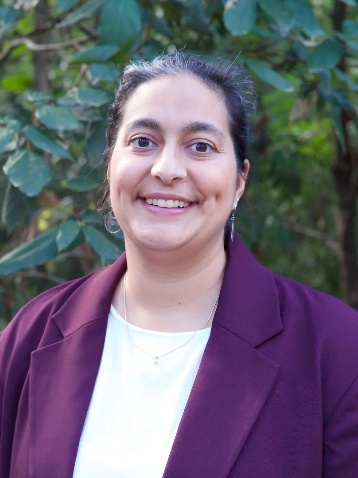 A woman with dark hair pulled back, smiling, wearing a white shirt, purple blazer, and jewelry, standing outdoors with green foliage in the background.