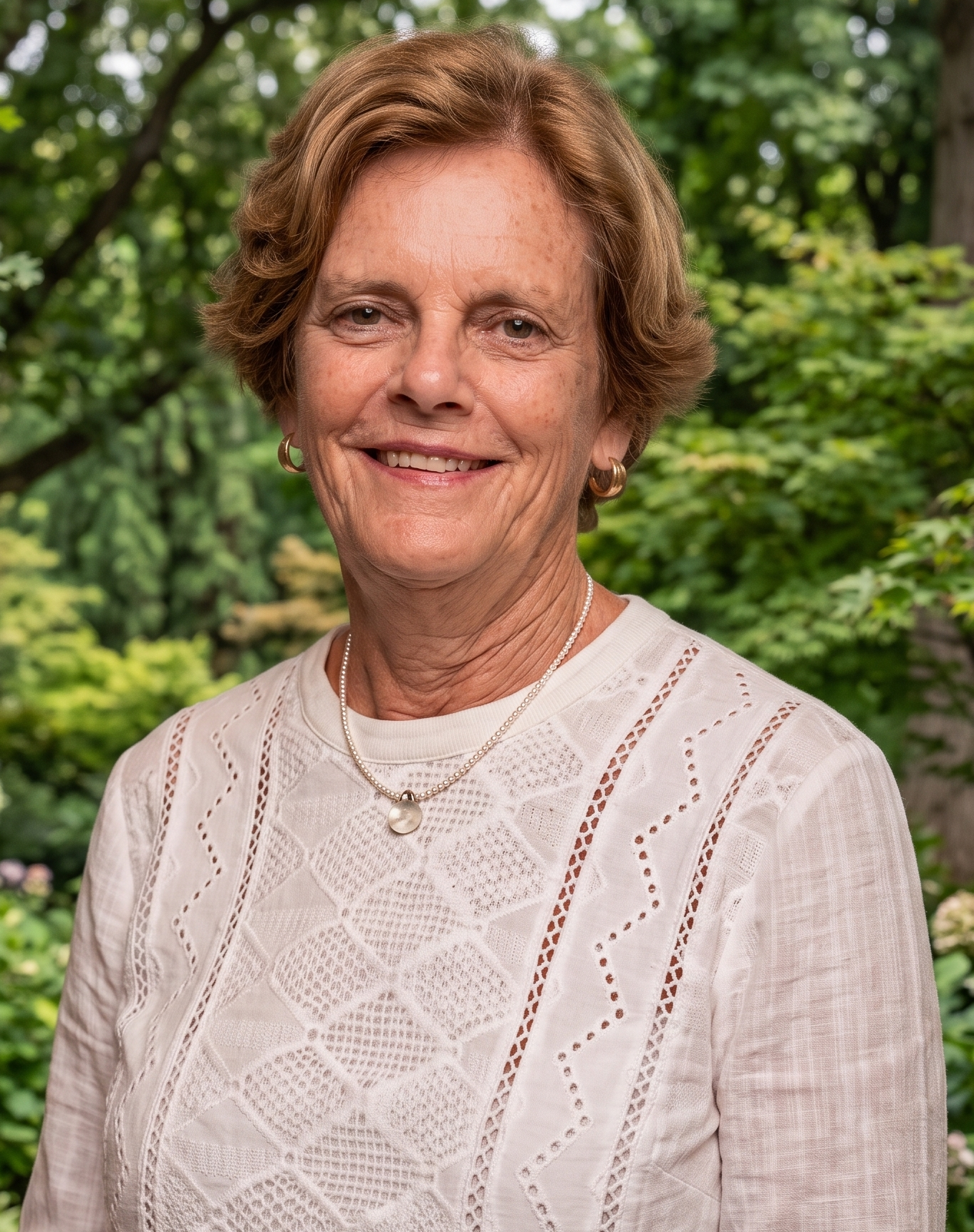 Portrait of an elderly woman with short, wavy light brown hair, wearing gold hoop earrings, a pearl necklace with a pendant, and a white, embroidered blouse, standing outdoors with green trees and foliage in the background.