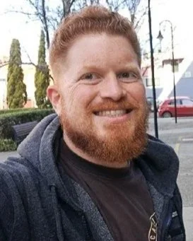 A man with red hair and a beard smiling outdoors with trees, street lamps, and parked cars in the background.