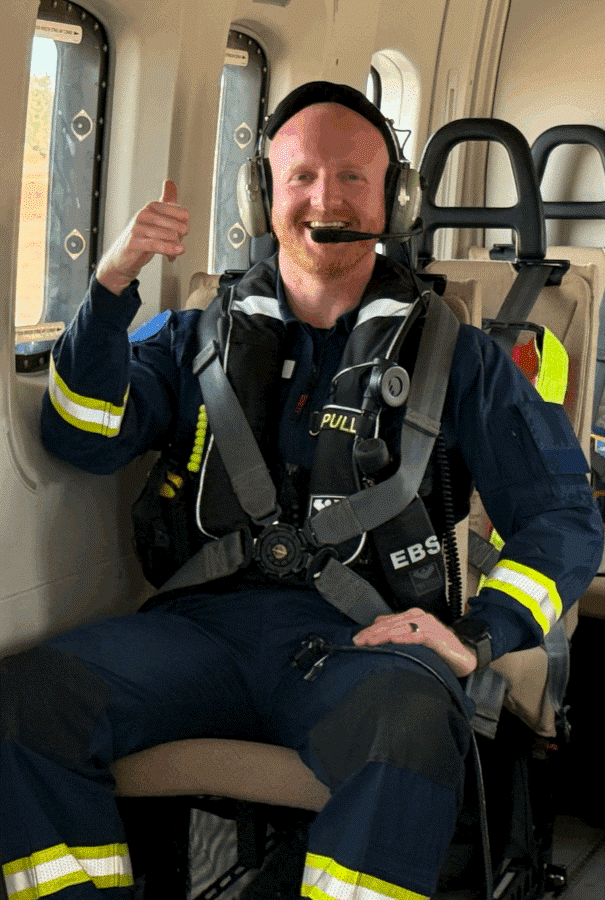 A smiling firefighter wearing a headset, sitting inside an aircraft, giving a thumbs-up, with a window showing trees outside.