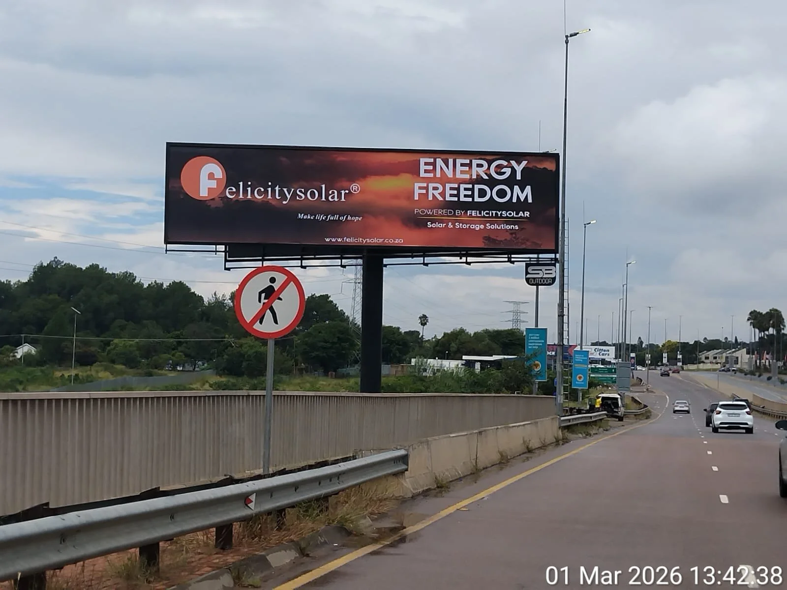 A billboard on a highway advertising Felicity Solar with the slogans 'Energy Freedom' and 'Make life full of hope'. There is a no pedestrian crossing sign in front of the billboard. The sky is cloudy, and there are cars on the road.