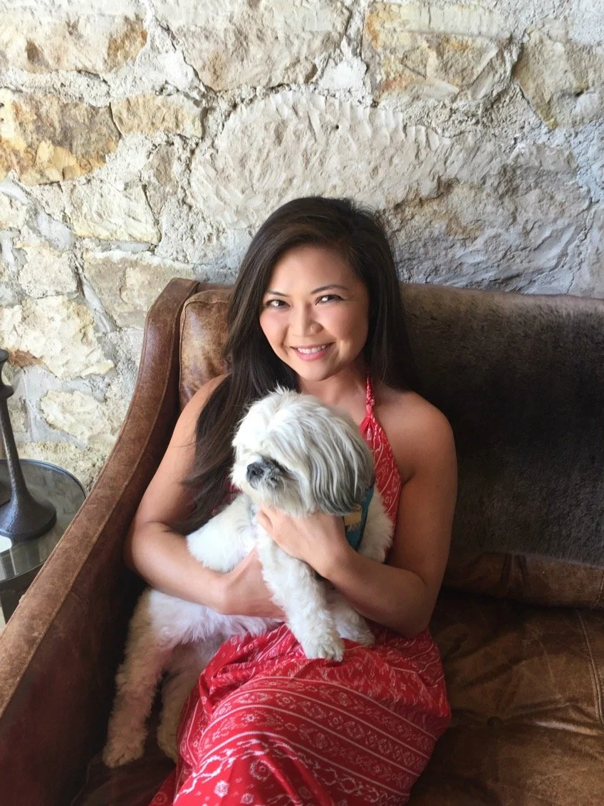 A woman in a red dress sitting on a brown sofa, holding a small white and gray dog, smiling and winking at the camera, with a stone wall in the background.