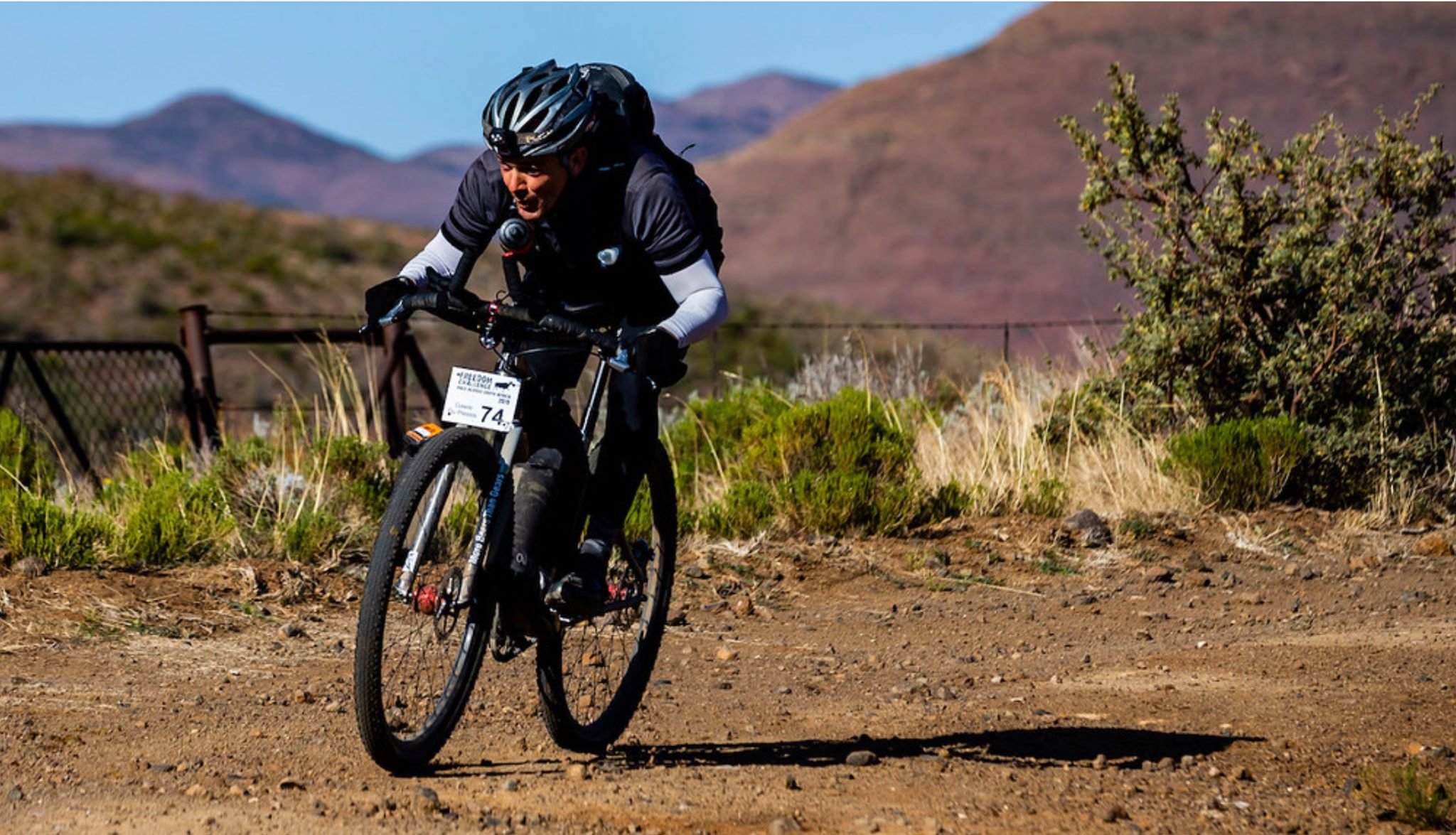 A person riding a mountain bike on a dirt trail in a desert landscape with mountains in the background.
