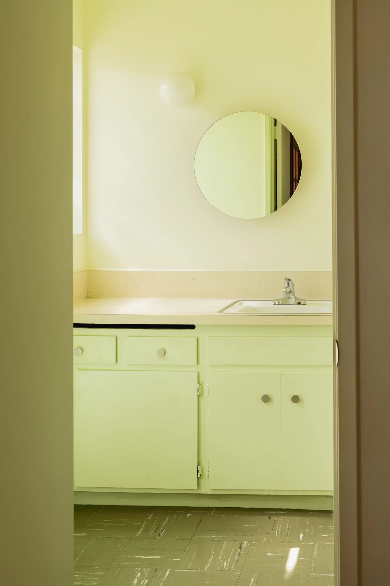 A bathroom with yellow cabinetry, a sink with a chrome fixture, a round mirror, and yellow walls, illuminated by natural light.