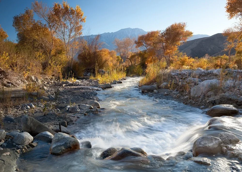 A mountain stream flowing through a landscape with autumn-colored trees and mountains in the background.