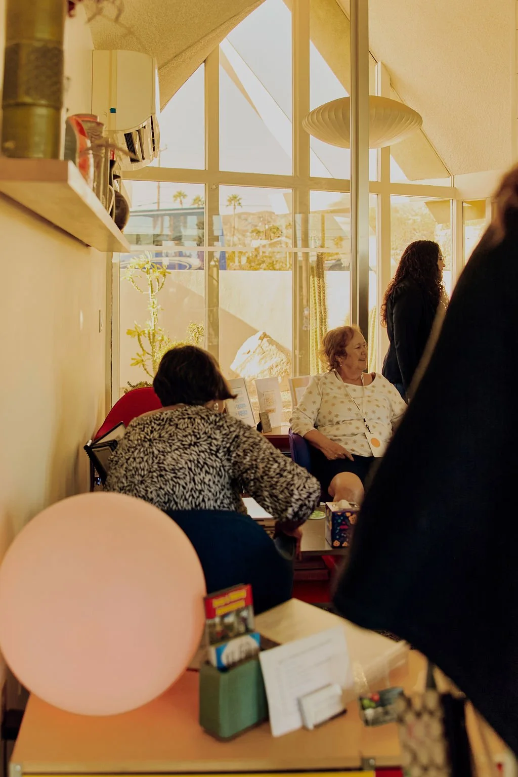 Interior of a well-lit room with large windows showing palm trees outside, where several women are seated and standing, engaged in conversation.