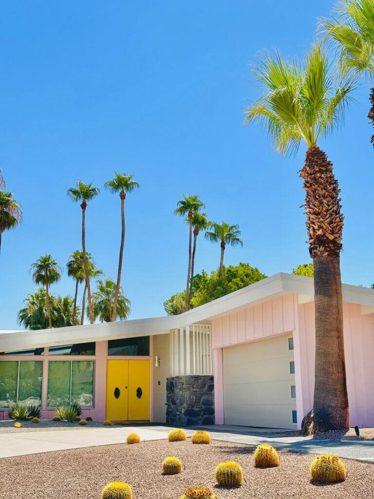 A mid-century modern house painted pink with a yellow door, set in a desert landscape with palm trees and bushes, under a clear blue sky.