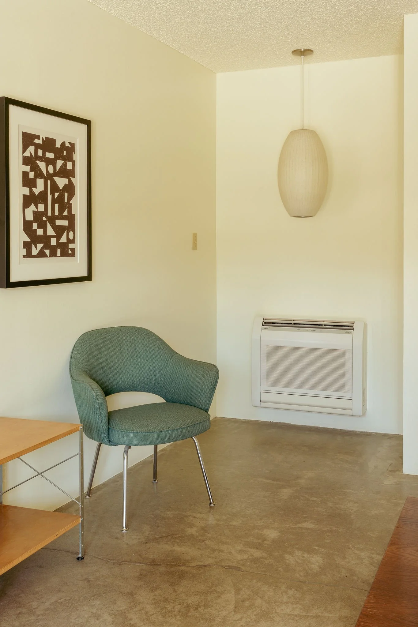 A green upholstered chair next to a wooden side table on a beige polished concrete floor, in a room with minimal decor. There is a framed abstract artwork with black and white shapes hanging on a light yellow wall, a hanging pendant lamp, and an air conditioning vent on the wall.