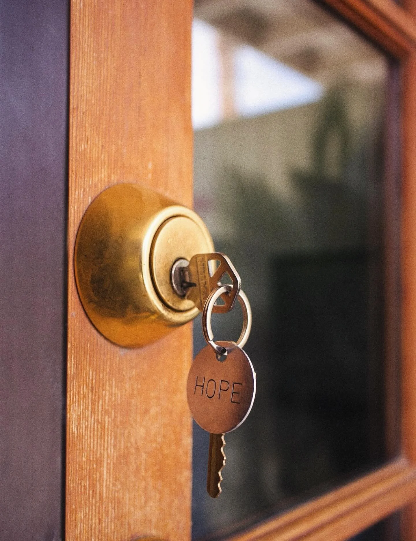 Close-up of a brass door lock with a key inserted, hanging on a wooden door. Attached to the key is a round metal tag with the word 'HOPE' engraved on it.