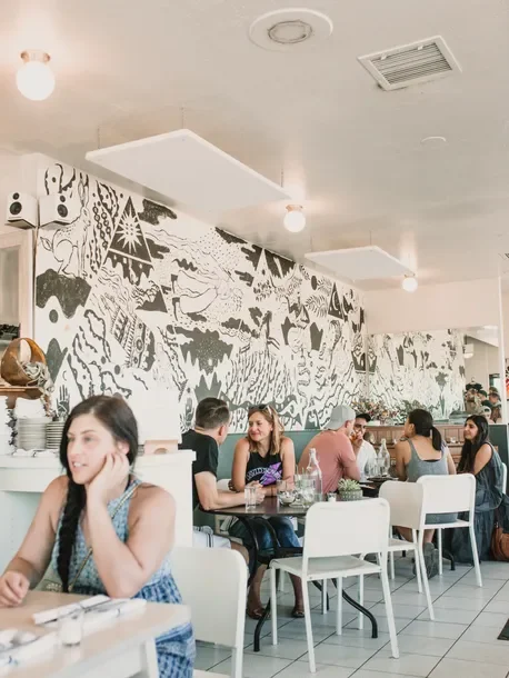 Cafe interior with white walls, black and white mural art, ceiling lights, and people dining at tables.