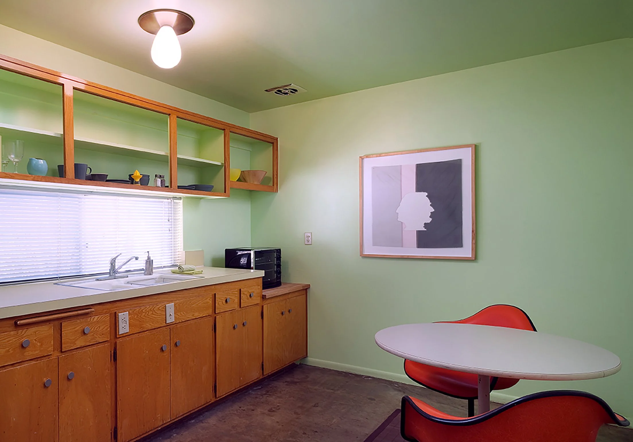 A kitchen with light green walls, wooden cabinets, a window with blinds, a wall art piece, a table with red chairs, and a ceiling light.