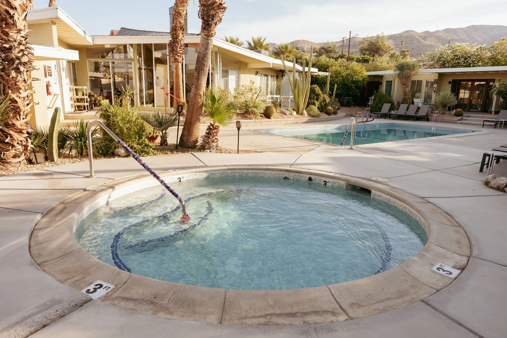 A resort or apartment complex pool area with a hot tub in the foreground, a swimming pool in the middle, and lounge chairs around the pools. Desert plants and palm trees decorate the area, with mountains visible in the background.