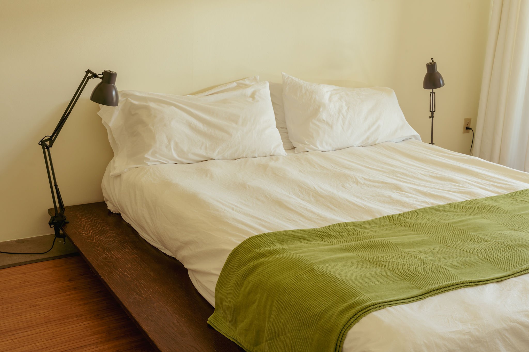 A neatly made bed with white pillows, white sheets, and a green quilt at the foot. Two black adjustable reading lamps are mounted on the wall above the bed, each with a power cord hanging down.