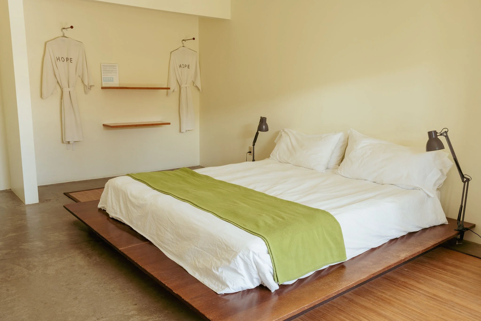 A minimalist bedroom with a low wooden bed frame, a white bed with white pillows, a green blanket, and two black adjustable reading lamps on either side. There are two cream-colored robes hanging on hooks on a cream wall, and small wooden shelves with a sign between them.
