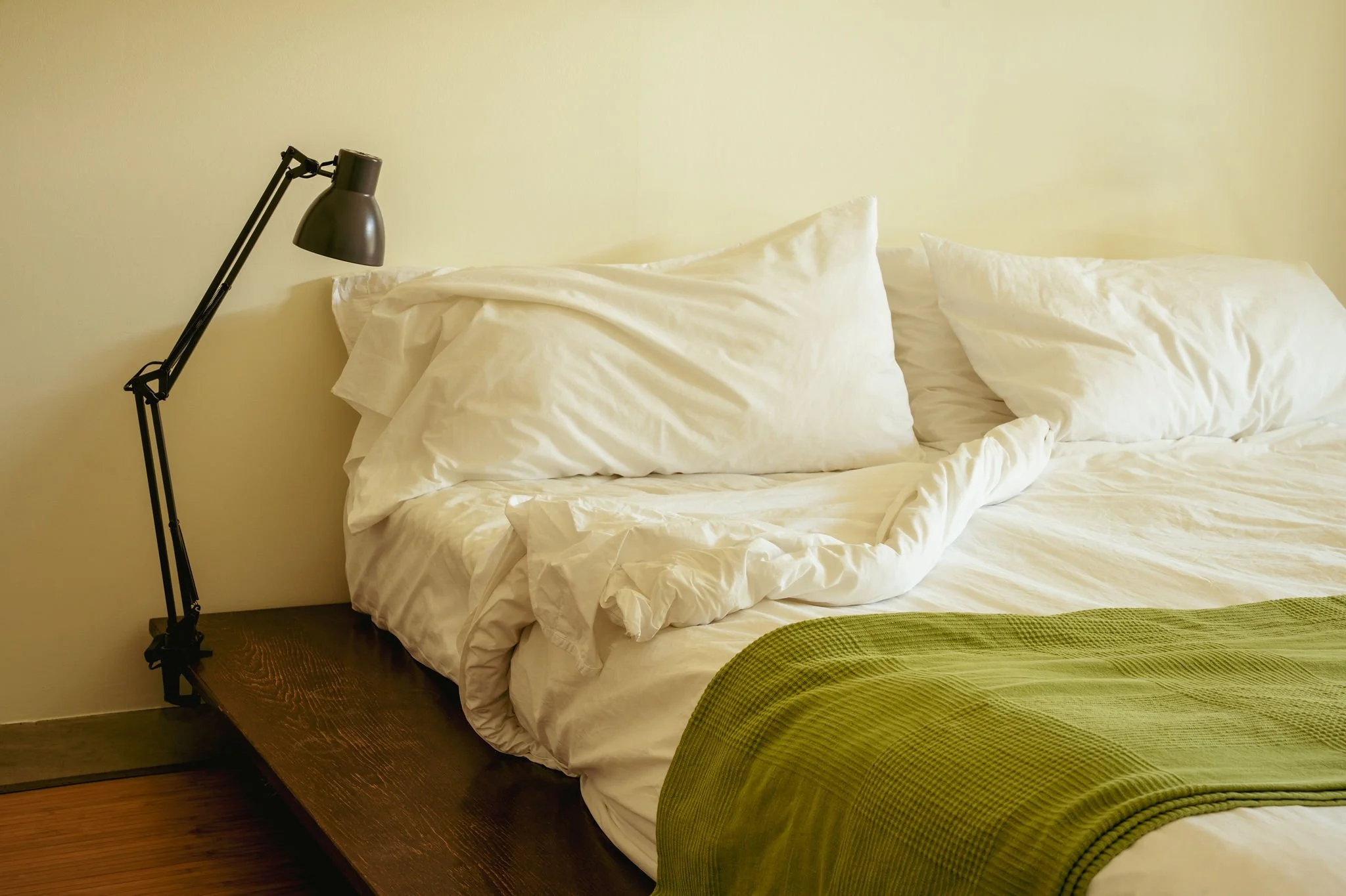 A neatly made bed with white sheets and pillows, a black adjustable reading lamp on a bedside table, and a green quilted blanket at the foot of the bed, against a plain yellow wall.