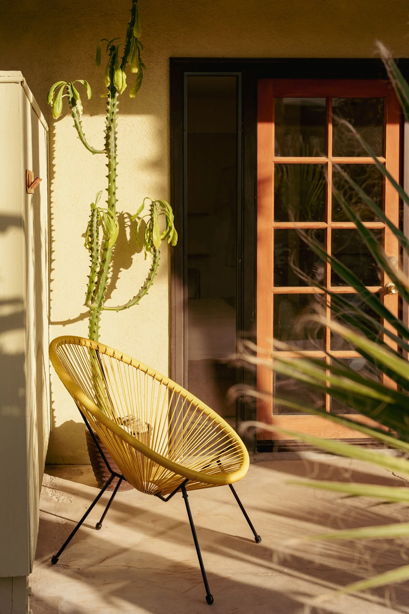 A yellow, woven, modern-style chair with black metal legs on a light-colored floor, near a sliding glass door with a wooden frame, next to a tall cactus plant and some greenery, in an outdoor patio area during daytime.