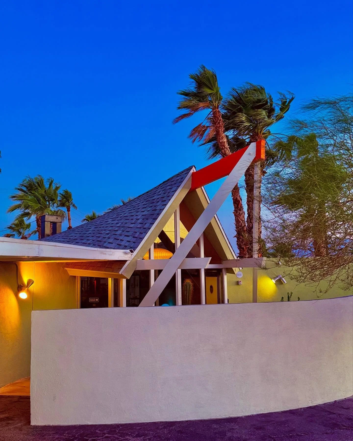 A building with a yellow exterior wall and a sloped roof, illuminated by yellow lights. Tall palm trees and a clear blue sky are visible in the background.