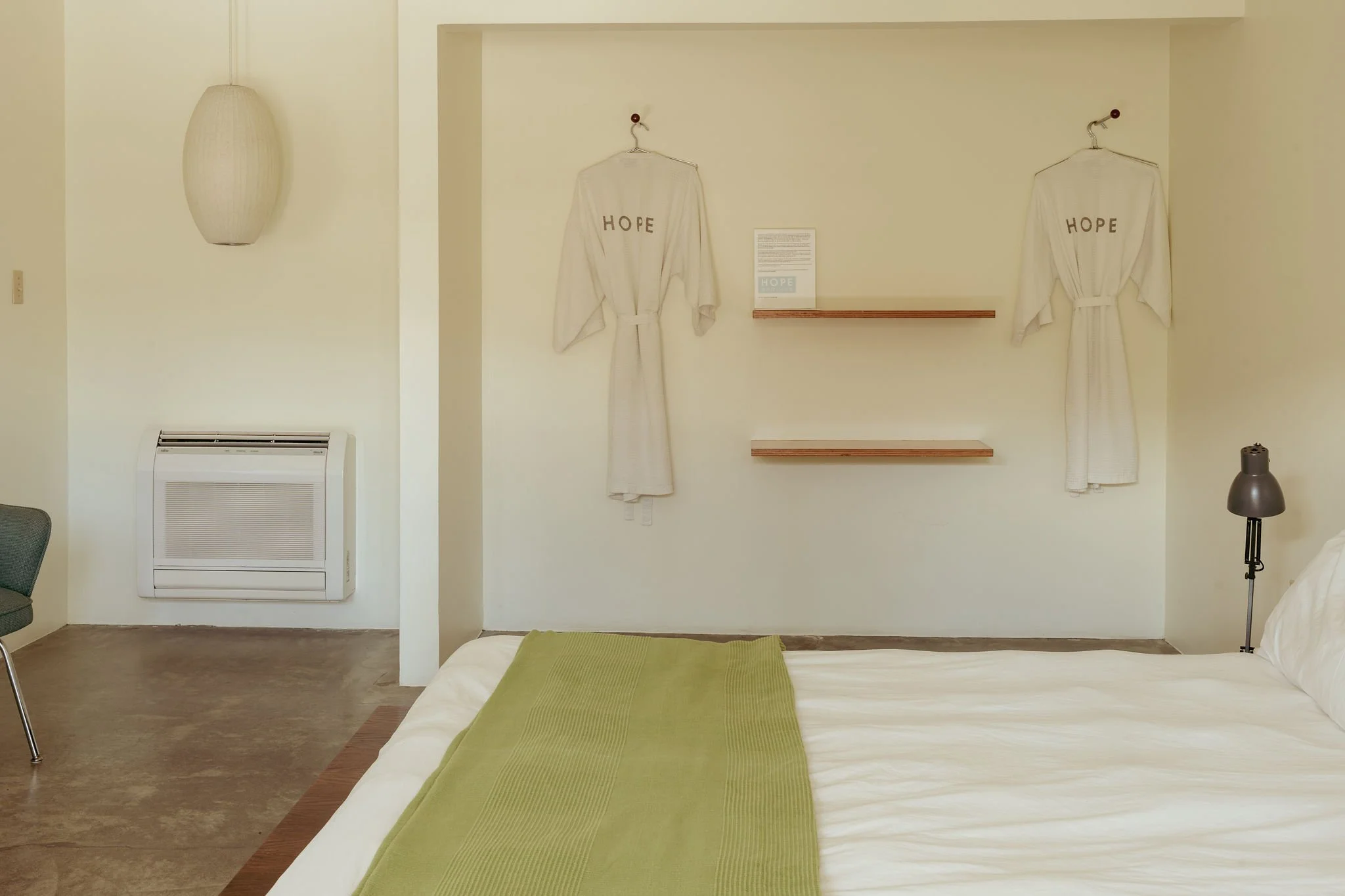 A hotel room with a bed covered in white sheets and a green runner, beige walls, a wall-mounted lamp, two white robes with 'HOPE' embroidered on them hanging on hooks, a small wooden shelf, a framed informational sign, and a black floor lamp.